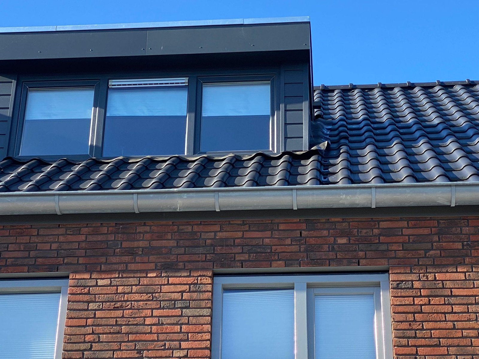 Brede antraciet dakkapel op een bakstenen woning met rolluiken