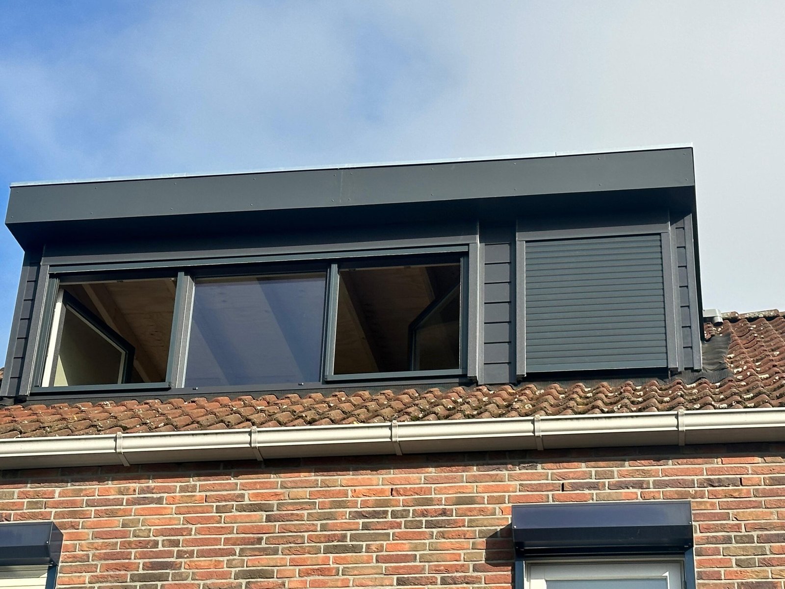 Grijze dakkapel met ingebouwde rolluiken op een woning met rode baksteen